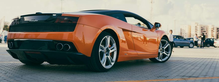 Orange Lamborghini sports car parked in an outdoor lot with other vehicles and people in the background.