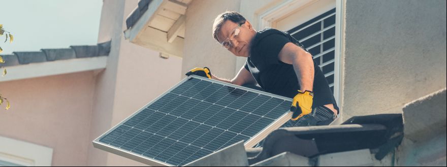 Photo of someone in work gloves and protective glasses on the roof of a home holding a solar panel.