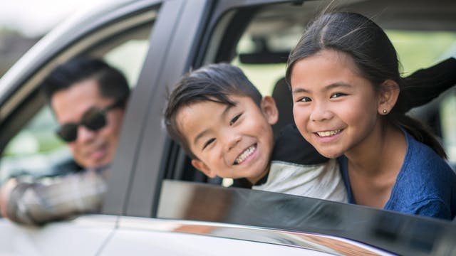 Man with sunglasses looking out of the front window of a car, kids with a smile looking out the back window.