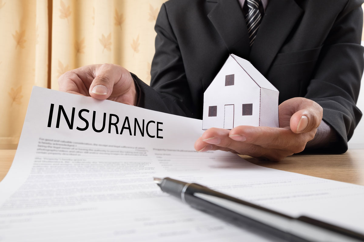 A hold on the right is holding a model of a house and the hand on the left holds up a page that says "Insurance"