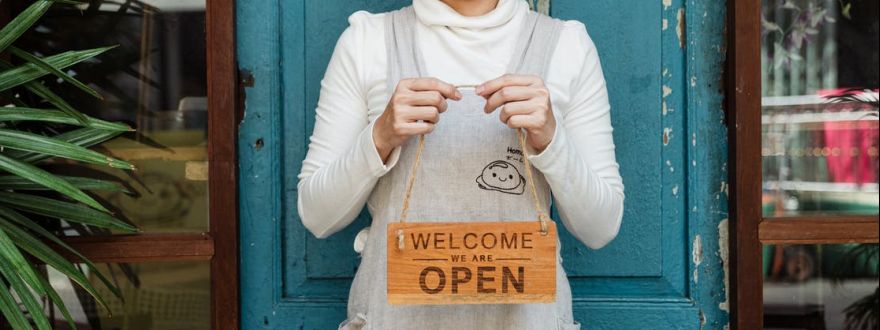 female cafeteria owner in apron demonstrating cardboard signboard while standing near blue shabby door and windows after starting own business and looking at camera