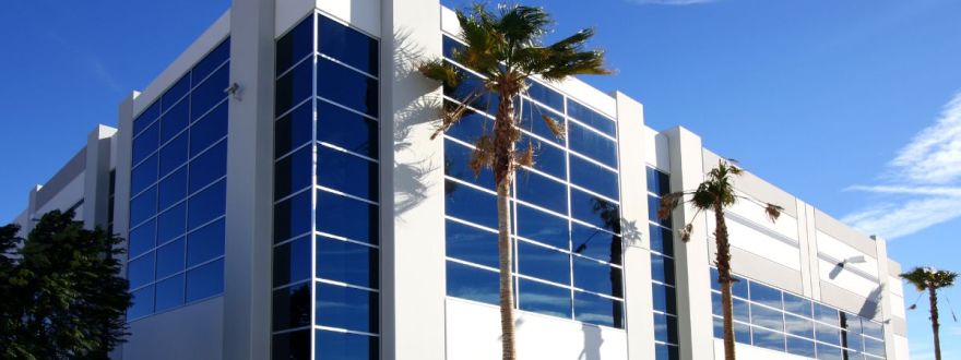 A modern commercial property building with glass windows and palm trees under a clear blue sky.
