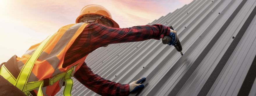 A worker in a safety vest and helmet using a drill to install metal roofing during sunset.