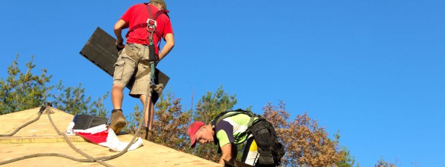 Two workers in safety gear installing a roof under a clear blue sky with trees in the background.