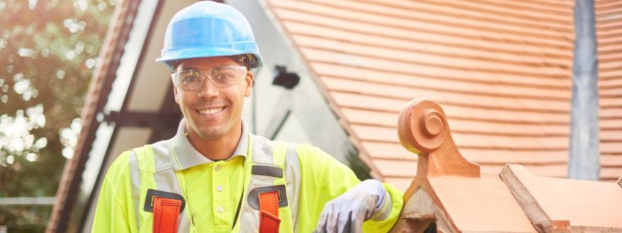 A smiling roofer worker in a blue hardhat and yellow safety vest standing in front of a roof under soft lighting.