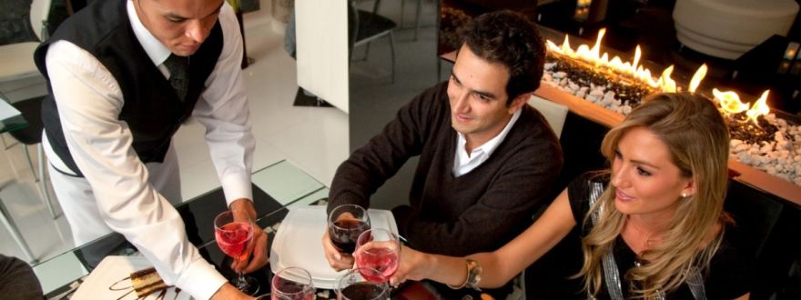 Restaurant server delivering drinks to a smiling couple seated at a modern, upscale dining table near a fireplace.