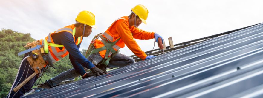 Two construction workers in safety gear installing a metal roof under a bright sky.