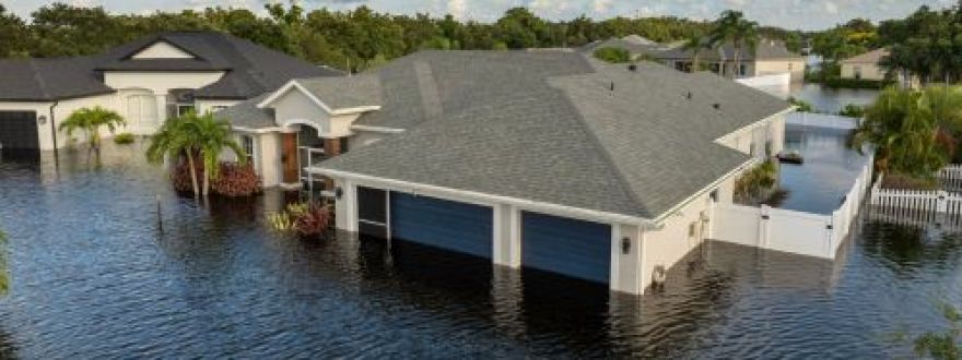 Aerial view of a modern suburban house with a two-car garage surrounded by dark floodwaters under a cloudy blue sky.