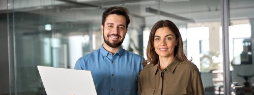A smiling man holding a laptop and a smiling woman stand side-by-side in a modern office with glass walls.