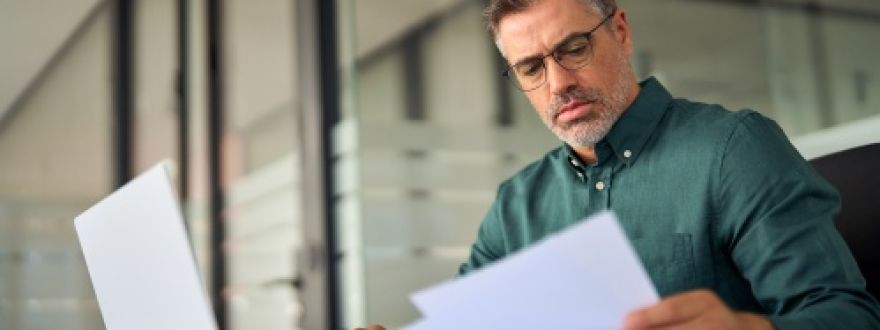 A man with glasses and a beard reviews documents while sitting in front of a laptop in an office.