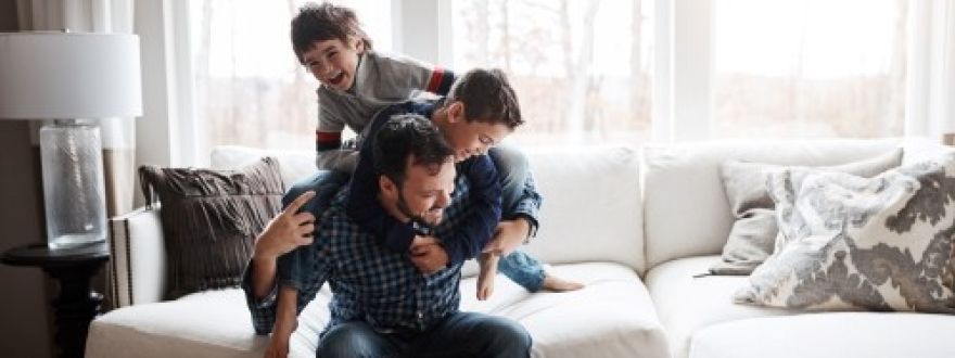 A father laughs while giving a piggyback ride to one son as another son sits beside them on a white couch in a sunlit room.