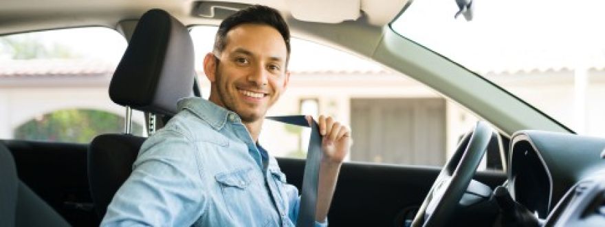 A smiling man in a denim shirt sits in a car's driver seat, looking at the camera while putting on his seatbelt.