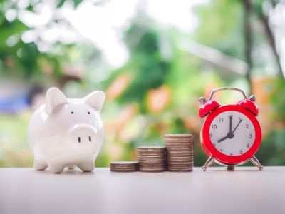 White piggy bank next to three stacks of coins and a red alarm clock, on a table with a blurred green background.