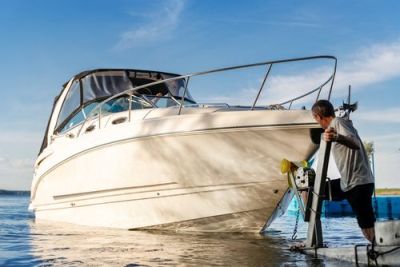 Man uses a trailer winch to launch a white cabin cruiser boat into the water on a sunny day.