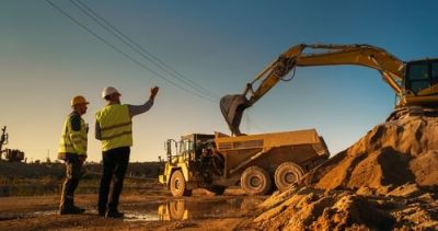 Two workers in safety gear watch an excavator load a dump truck at a construction site during sunset.