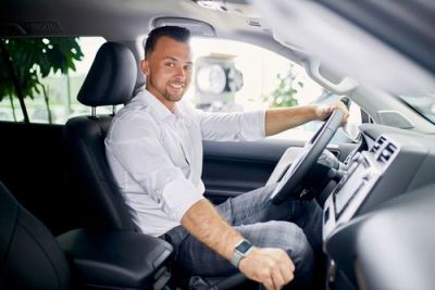 Man in a white shirt smiles while sitting in the driver's seat of a car, one hand on the steering wheel.