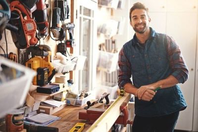 Smiling contractor leans on workbench in his workshop, holding a pencil, with tools on the wall behind.