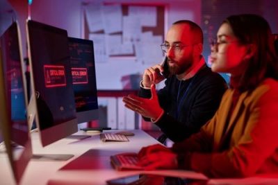 IT team responds to critical computer error; man on phone, woman typing, red alert light reflects on faces.