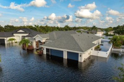 Aerial view of a modern suburban house with a two-car garage surrounded by dark floodwaters under a cloudy blue sky.