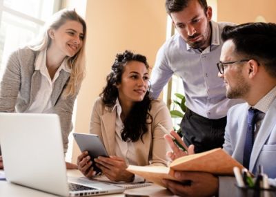 Four young professionals collaborating around an office desk with laptops and a tablet.