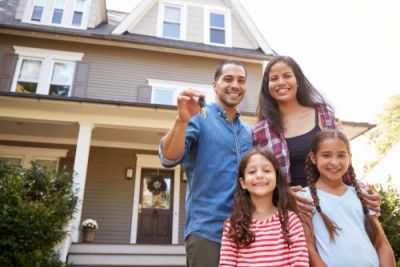 Smiling family of four with two young daughters standing outside their two-story home; the father holds up a set of keys.