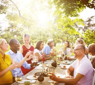 An outdoor gathering, a diverse group sits at a long table, enjoying food and drinks under sunny trees.