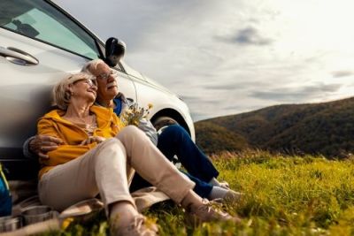 Senior couple relaxing together, leaning against their car in a grassy field with mountains in the background.