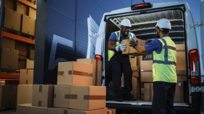 Two diverse workers in hard hats smile while loading boxes into a delivery van in a warehouse.