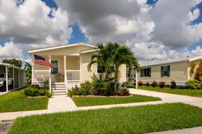 Yellow manufactured home with a porch, American flag, palm tree, and neat lawn under a partly cloudy sky.