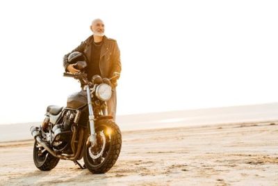 Smiling mature man with a beard, wearing a leather jacket, standing with his motorcycle in a bright, sandy landscape.