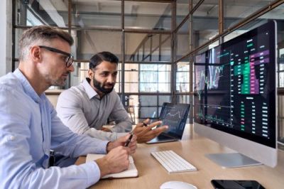 Two male financial professionals analyze trading charts on computer screens in modern office.