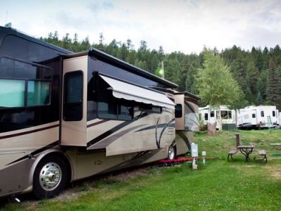 Large tan and brown RV with slide-outs and an awning extended, parked in a grassy campground with trees.
