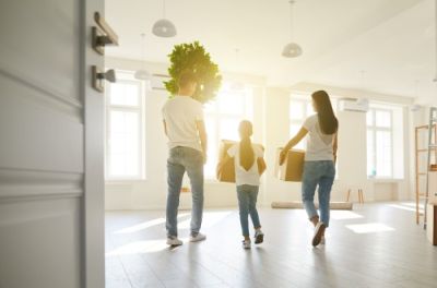 Back view of a family of three carrying boxes and a plant into a bright, empty new apartment, seen from the doorway.