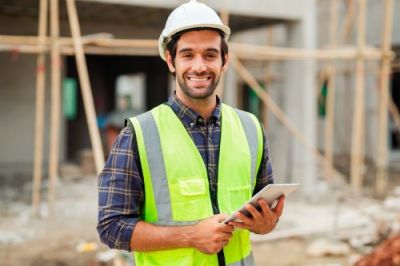 Smiling construction engineer in hard hat & safety vest holding tablet on job site.